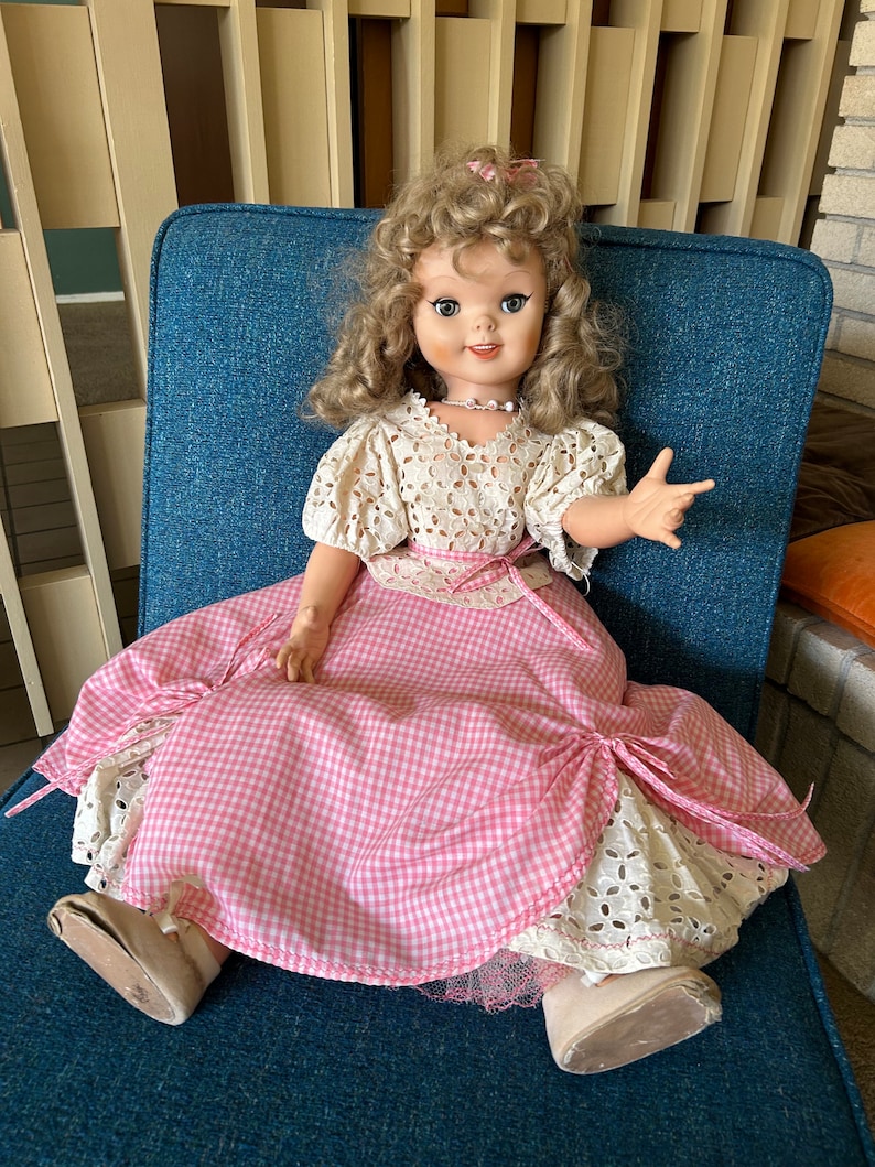 May include: A vintage doll with blonde curly hair, wearing a white lace top and a pink gingham dress. The doll is seated on a blue chair, with a pearl necklace and a pink bow in her hair. The doll's shoes are beige.