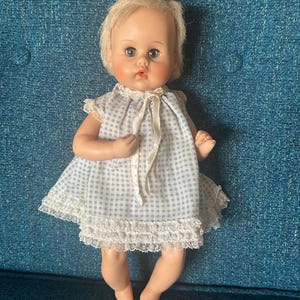 May include: A vintage baby doll with blonde hair and blue eyes. The doll wears a blue and white checkered dress with lace trim and a white ribbon. The doll is sitting on a blue surface.