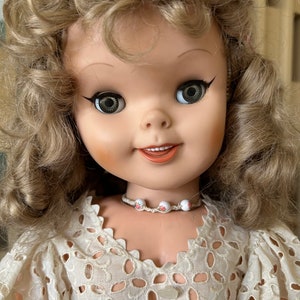 May include: A vintage doll with blonde curly hair and large, expressive eyes. The doll wears a white, eyelet lace dress and a small beaded necklace with floral accents. The doll's face is painted with rosy cheeks and a subtle smile.