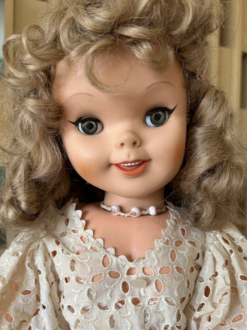 May include: A vintage doll with blonde curly hair and large, expressive eyes. The doll wears a white, eyelet lace dress and a small beaded necklace with floral accents. The doll's face is painted with rosy cheeks and a subtle smile.