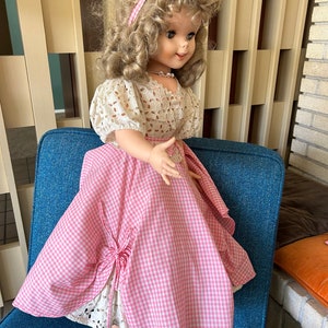 May include: A vintage doll with blonde curly hair, wearing a pink and white gingham dress with a white lace overlay. The doll has a pink headband and a pearl necklace. The doll is seated on a blue chair.