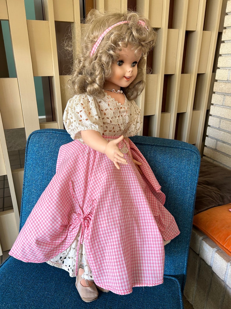 May include: A vintage doll with blonde curly hair, wearing a pink and white gingham dress with a white lace overlay. The doll has a pink headband and a pearl necklace. The doll is seated on a blue chair.