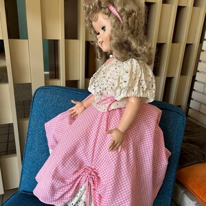 May include: A vintage doll with blonde curly hair, wearing a pink and white gingham dress with a white lace overlay. The doll is seated on a blue chair. The doll's dress has a bow detail.