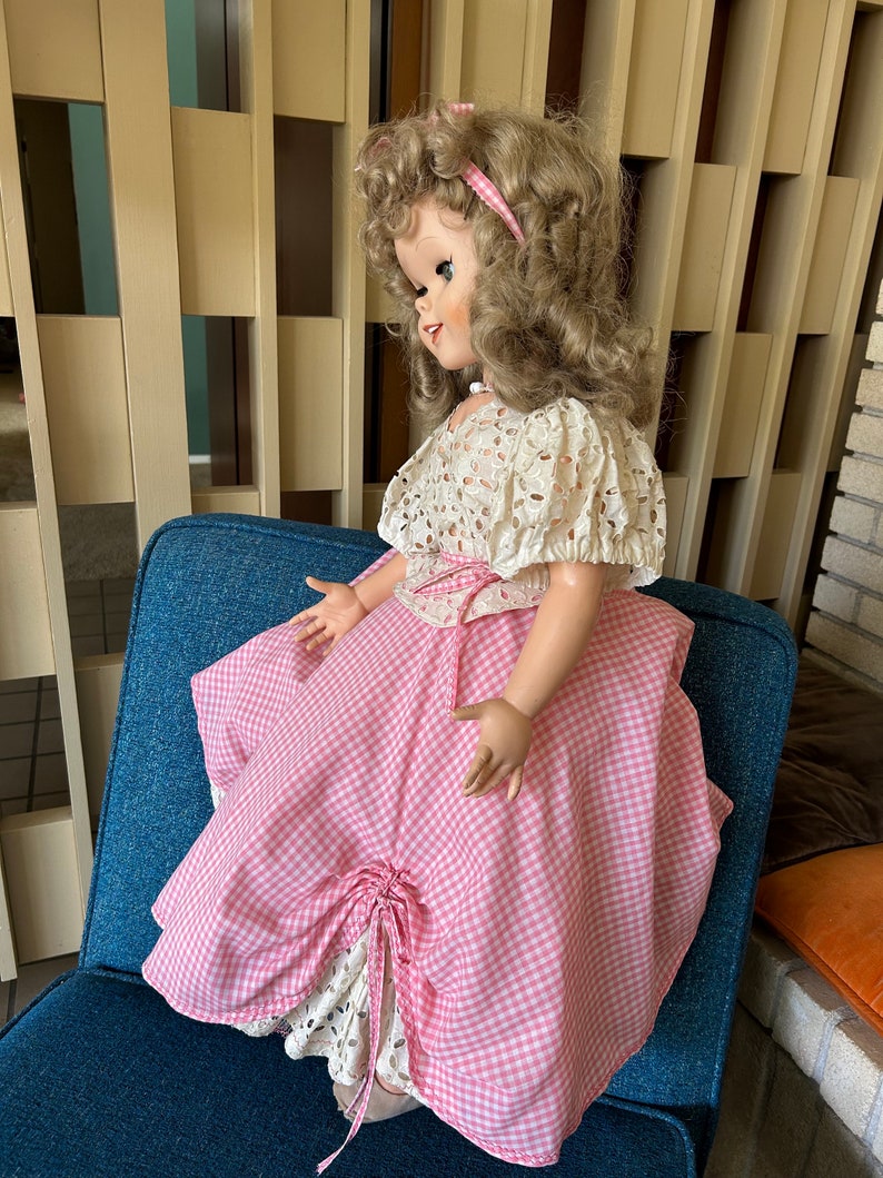 May include: A vintage doll with blonde curly hair, wearing a pink and white gingham dress with a white lace overlay. The doll is seated on a blue chair. The doll's dress has a bow detail.