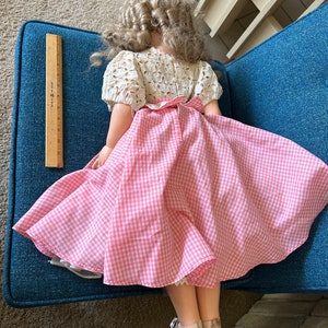 May include: A vintage doll with blonde curly hair, wearing a white eyelet top and a pink and white gingham skirt. The doll's skirt is flared, and it has white shoes. A wooden ruler is next to the doll.