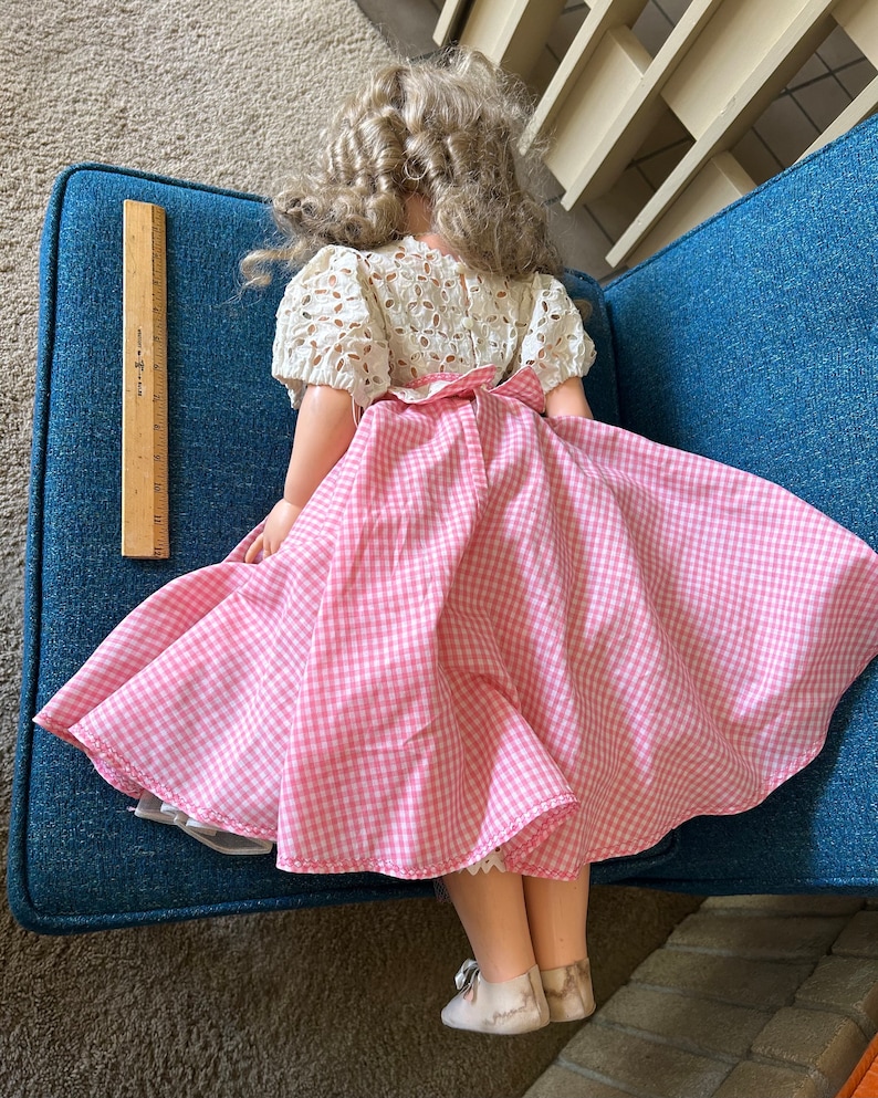 May include: A vintage doll with blonde curly hair, wearing a white eyelet top and a pink and white gingham skirt. The doll's skirt is flared, and it has white shoes. A wooden ruler is next to the doll.