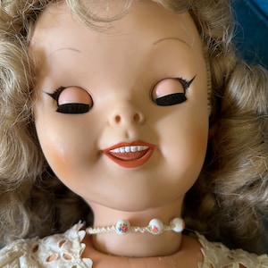 May include: A vintage doll with closed eyes, rosy cheeks, and a slight smile. The doll has blonde, curly hair and wears a cream-colored lace dress. A delicate necklace with floral beads adorns the doll's neck.
