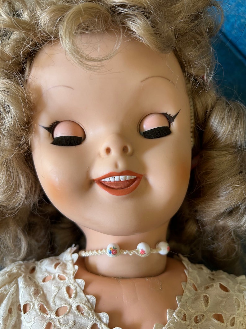 May include: A vintage doll with closed eyes, rosy cheeks, and a slight smile. The doll has blonde, curly hair and wears a cream-colored lace dress. A delicate necklace with floral beads adorns the doll's neck.