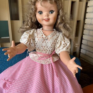 May include: A vintage doll with blonde curly hair, wearing a white eyelet top and a pink and white gingham skirt. The doll has large blue eyes and is posed with open arms. A pink ribbon accents the waist.