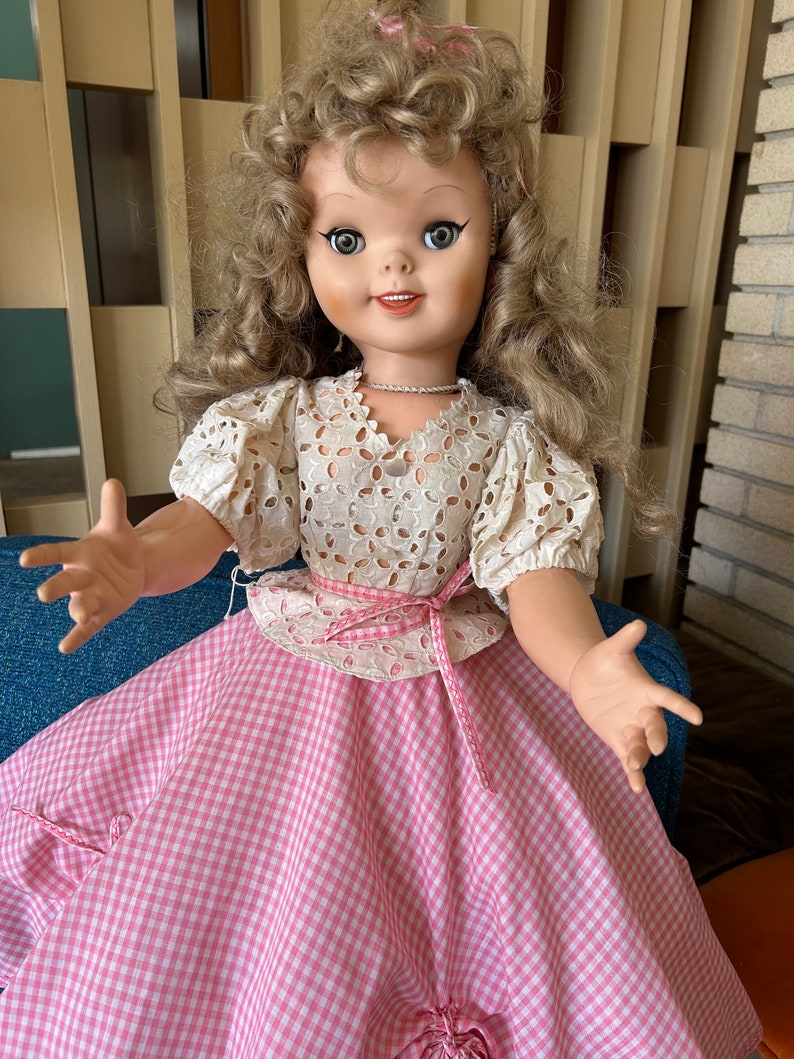 May include: A vintage doll with blonde curly hair, wearing a white eyelet top and a pink and white gingham skirt. The doll has large blue eyes and is posed with open arms. A pink ribbon accents the waist.