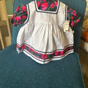 May include: A white dress with a blue and red floral trim. The dress has a white bodice and a white skirt with a blue and red floral trim. The dress is on a blue chair.