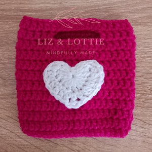 May include: A small pink crocheted bag with a white crocheted heart on the front. The bag has a handle and says "Liz & Lottie Mindfully Made".