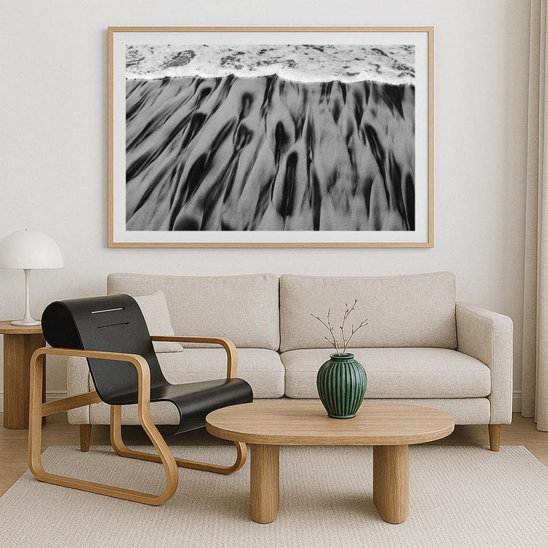 May include: A framed black and white photograph of a beach scene. The image captures the textured patterns of the sand and the foamy edge of the ocean waves. The photograph is displayed above a beige sofa, a black chair, and a wooden coffee table.