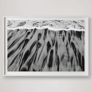 Puede incluir: Fotograf&iacute;a en blanco y negro enmarcada de una escena de playa. La imagen captura la textura de la arena mojada con patrones oscuros y fluidos, y el borde espumoso de las olas del oc&eacute;ano. La fotograf&iacute;a se muestra en un marco blanco.
