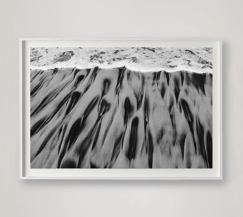 Puede incluir: Fotograf&iacute;a en blanco y negro enmarcada de una escena de playa. La imagen captura la textura de la arena mojada con patrones oscuros y fluidos, y el borde espumoso de las olas del oc&eacute;ano. La fotograf&iacute;a se muestra en un marco blanco.
