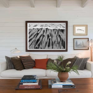 Puede incluir: Un sal&oacute;n con un sof&aacute; blanco y cojines en tonos marrones y naranjas. Sobre el sof&aacute;, una gran fotograf&iacute;a en blanco y negro enmarcada de una escena de playa. Una mesa de centro de madera exhibe libros y una planta en maceta.