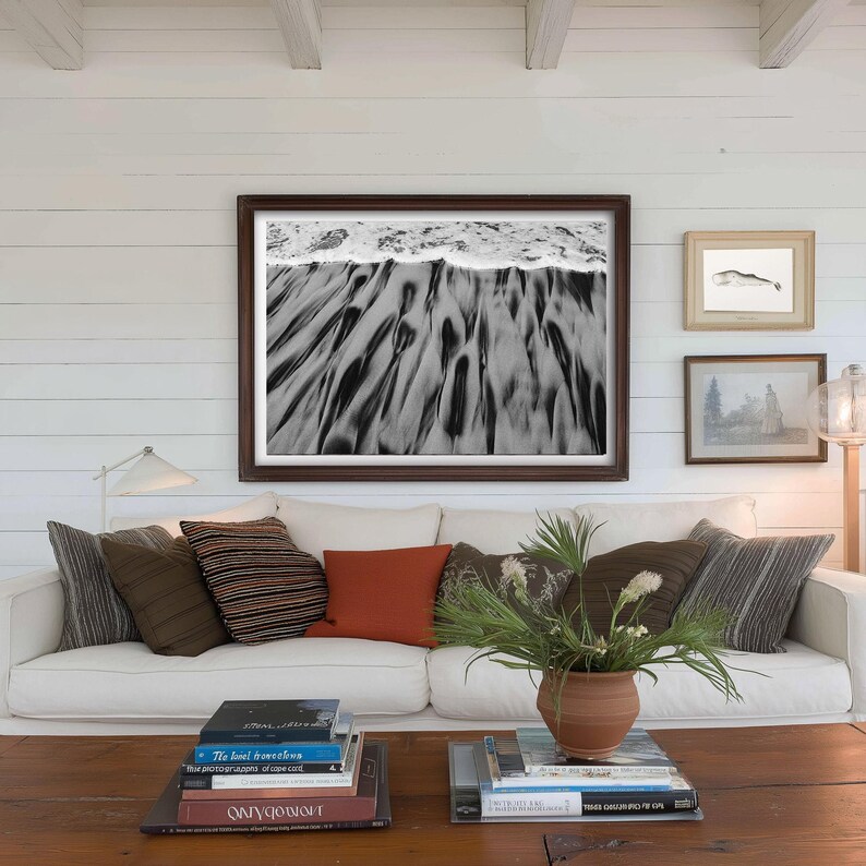 Puede incluir: Un sal&oacute;n con un sof&aacute; blanco y cojines en tonos marrones y naranjas. Sobre el sof&aacute;, una gran fotograf&iacute;a en blanco y negro enmarcada de una escena de playa. Una mesa de centro de madera exhibe libros y una planta en maceta.