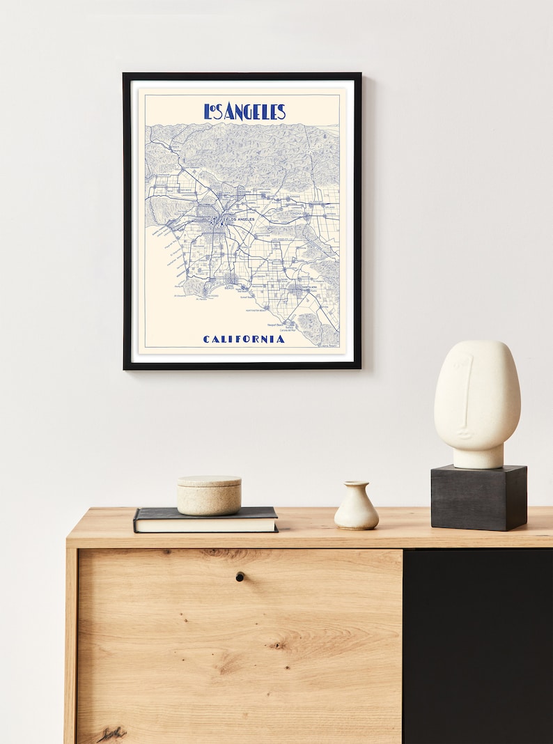 Pu&ograve; includere: Una mappa incorniciata di Los Angeles, California, con linee blu che rappresentano strade e autostrade. La mappa &egrave; su una parete bianca sopra una credenza in legno con una scultura in bianco e nero, un piccolo vaso bianco e un libro.
