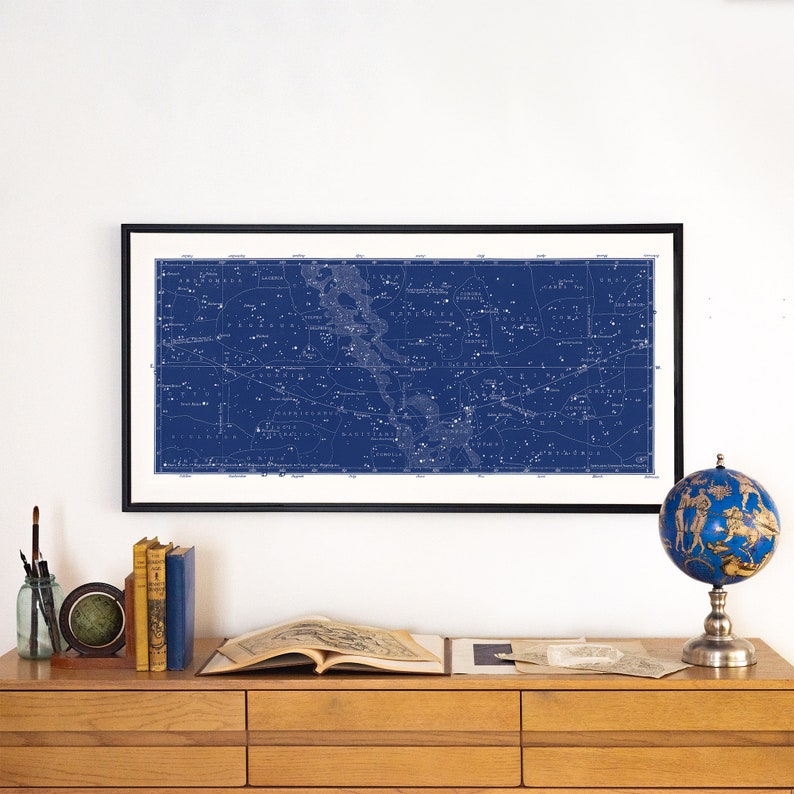 Puede incluir: Un mapa de estrellas azul y blanco enmarcado con las constelaciones y sus nombres impresos en &eacute;l. El mapa est&aacute; colgado en una pared blanca sobre un escritorio de madera con libros, un globo terr&aacute;queo y un libro abierto sobre el escritorio.