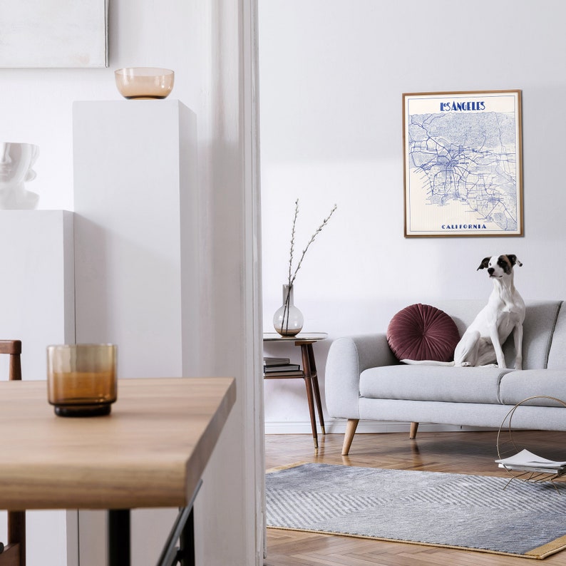 Pu&ograve; includere: Una stampa di mappa blu e bianca di Los Angeles, California, incorniciata in una cornice marrone, appesa a un muro bianco sopra un divano grigio con un cuscino di lancio bordeaux. Un cane bianco &egrave; seduto sul divano.