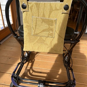 May include: A black walker with a yellow fabric bag attached to the back. The bag has a pocket and two buttons. The walker has two wheels on the front and two wheels on the back.
