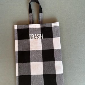 Puede incluir: Una bolsa de basura a cuadros blancos y negros con un asa negra. La palabra "TRASH" está impresa en letras blancas en la parte delantera. La bolsa es de tela.