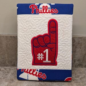 May include: A quilted fabric item featuring a red and blue #1 foam finger design. The word "Phillies" is printed in white on a blue background. The item has a white quilted center.