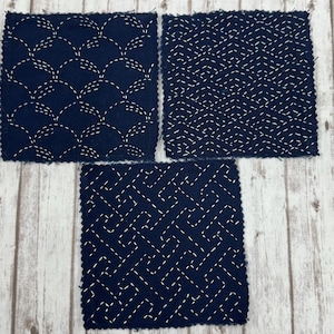 May include: Three square fabric patches in dark navy blue, each featuring distinct white stitched patterns. One patch displays a repeating wave design, another shows a geometric diamond grid, and the third features a complex interlocking maze pattern.