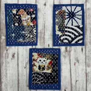 May include: Three rectangular fabric patches featuring blue borders and white sashiko-style stitching. The patches display various Japanese-inspired patterns, including a beckoning cat, floral motifs, geometric lines, and wave designs in navy, white, and gold.