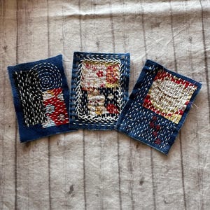 May include: Three small, square textile art pieces with intricate stitching. Each piece features a dark blue background with white and red thread accents. The designs include geometric patterns and abstract shapes, showcasing a handcrafted aesthetic.