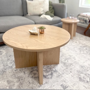 May include: A round wooden coffee table with a light wood finish. The table has four legs that are connected in the middle. There is a small potted succulent plant and a crystal on the table.
