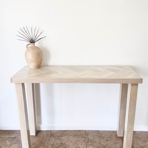 May include: A light wood console table with a herringbone pattern top and four legs. The table is in front of a white wall and has a small vase with dried branches on top.