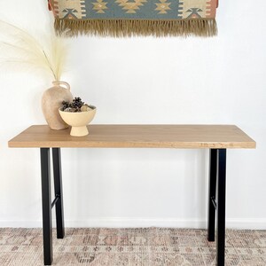 May include: A light wood console table with black metal legs. The table is sitting on a patterned rug. There is a vase with dried flowers and a bowl of decorative items on the table.