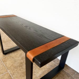 May include: A modern black bench with a rectangular top and black metal legs. The top is made of dark wood and features a brown leather accent on each end. The bench is approximately 48 inches long.