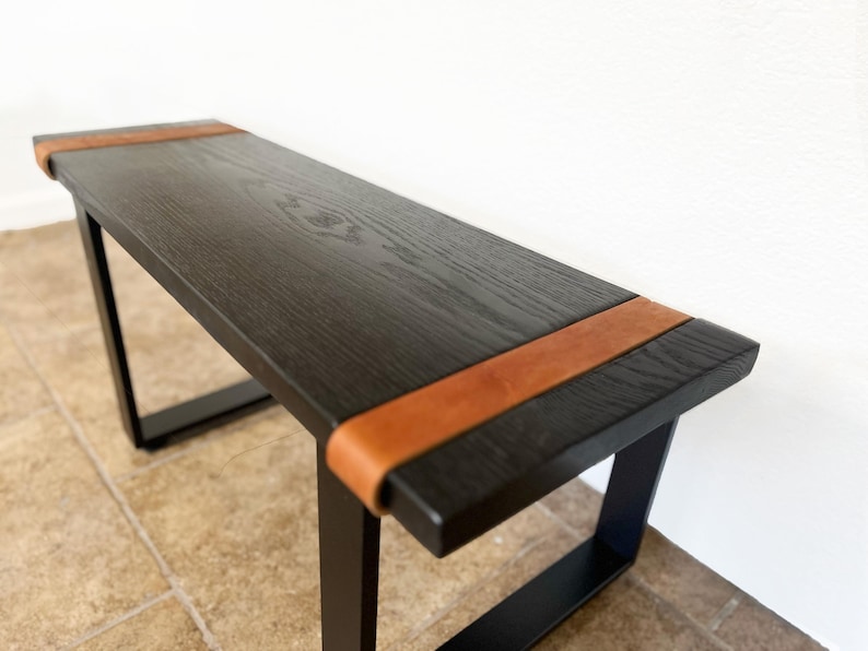 May include: A modern black bench with a rectangular top and black metal legs. The top is made of dark wood and features a brown leather accent on each end. The bench is approximately 48 inches long.
