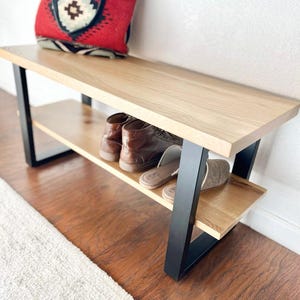 May include: A wooden bench with a light-colored top and a lower shelf, supported by black metal legs. The bench holds a pair of brown leather shoes and a pair of tan slippers. A red and patterned bag rests on the bench.