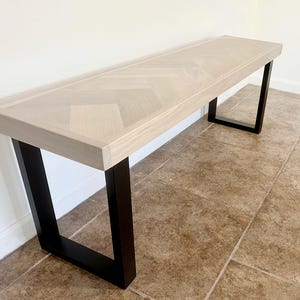 May include: A light-colored wooden bench with a rectangular top and black metal legs. The bench top features a herringbone pattern. The legs are a simple, modern design. The bench is suitable for a hallway or entryway.
