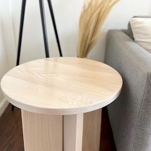 May include: A light-colored, round wooden side table. The table has a circular top and a rectangular base with vertical supports. The wood grain is visible. The table is next to a gray sofa and a tall black tripod lamp.