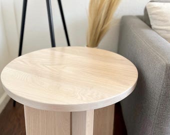 May include: A light-colored, round wooden side table. The table has a circular top and a rectangular base with vertical supports. The wood grain is visible. The table is next to a gray sofa and a tall black tripod lamp.