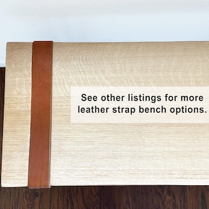 May include: A light-colored wooden bench with a brown leather strap detail. The bench has a rectangular shape and is made of light-colored wood. The leather strap is vertically placed on the side of the bench. Text on the bench reads: See other listings for more leather strap bench options.