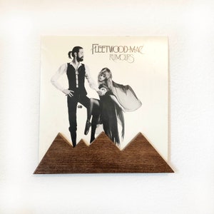 May include: A brown wooden record holder shaped like mountains holds a vinyl record with a black and white photo of Fleetwood Mac on the cover. The album title is "Rumours".