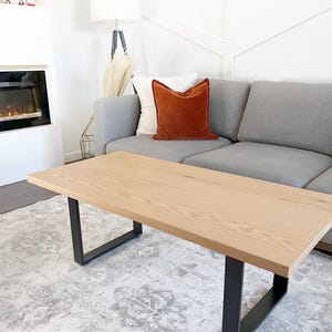 May include: A light wood coffee table with black metal legs sits on a gray and white patterned rug. The table is in a living room setting with a gray sofa and a floor lamp with a white shade.