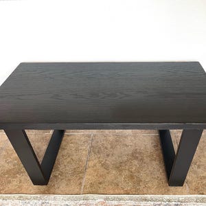 May include: A rectangular black wooden table with a dark finish. The table has a simple design with V-shaped legs. The table is approximately 48 inches long and 24 inches wide, suitable for various home decor styles.