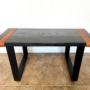 May include: A rectangular black wooden coffee table with angled black legs. The table features a brown leather strap detail on each end. The table is approximately 48 inches long, 20 inches wide, and 18 inches high.