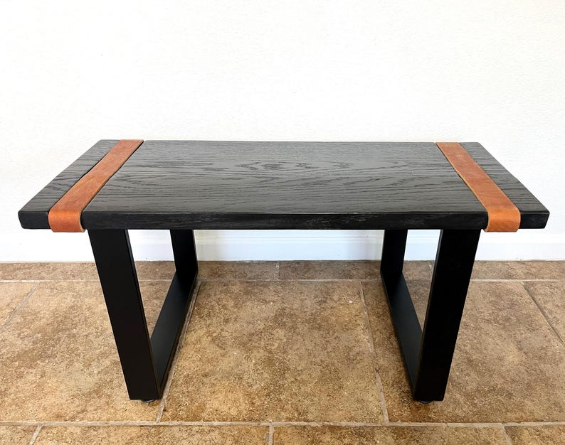 May include: A rectangular black wooden coffee table with angled black legs. The table features a brown leather strap detail on each end. The table is approximately 48 inches long, 20 inches wide, and 18 inches high.