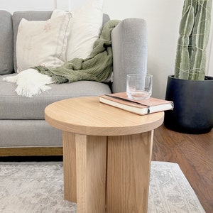 May include: A light wood round side table with four legs. The table has a book and a glass on top. The table is in front of a gray couch with a white throw blanket and a green throw blanket.