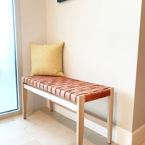 May include: A light wood bench with a woven leather seat and a yellow pillow. The bench is in a corner of a room with white walls and a framed picture on the wall.
