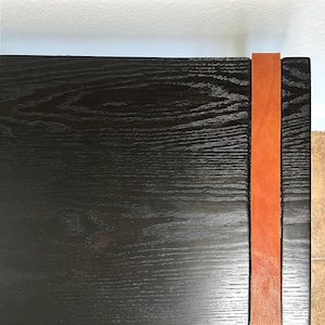 May include: A close-up of a black wooden table with a visible wood grain texture. A vertical orange leather strap is attached to the side of the table, adding a contrasting color and texture. The table's design is simple and modern.