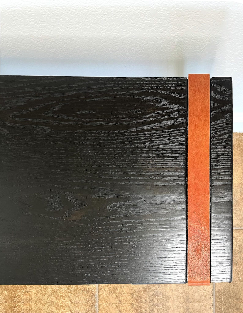 May include: A close-up of a black wooden table with a visible wood grain texture. A vertical orange leather strap is attached to the side of the table, adding a contrasting color and texture. The table's design is simple and modern.