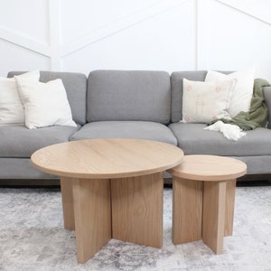 May include: Two wooden coffee tables with a gray sofa in the background. The larger table is round with a natural wood finish. The smaller table is square and also has a natural wood finish.